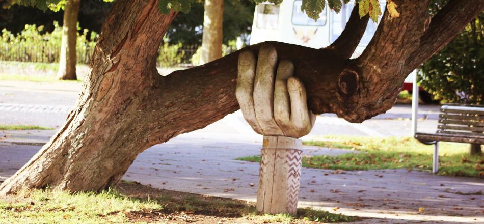 a carved wooden hand coming out of the ground is holding up the trunk of a tree